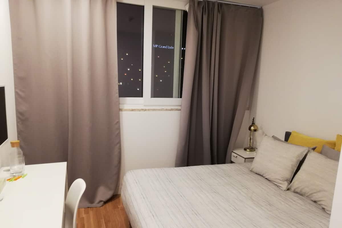 A hotel room with a bed, a desk, a chair, a nightstand, and a window with curtains.