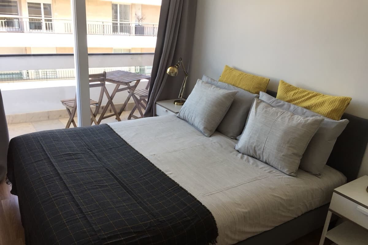 A bedroom with a bed, pillows, a nightstand, a lamp, and a view of a balcony with a table and chairs.