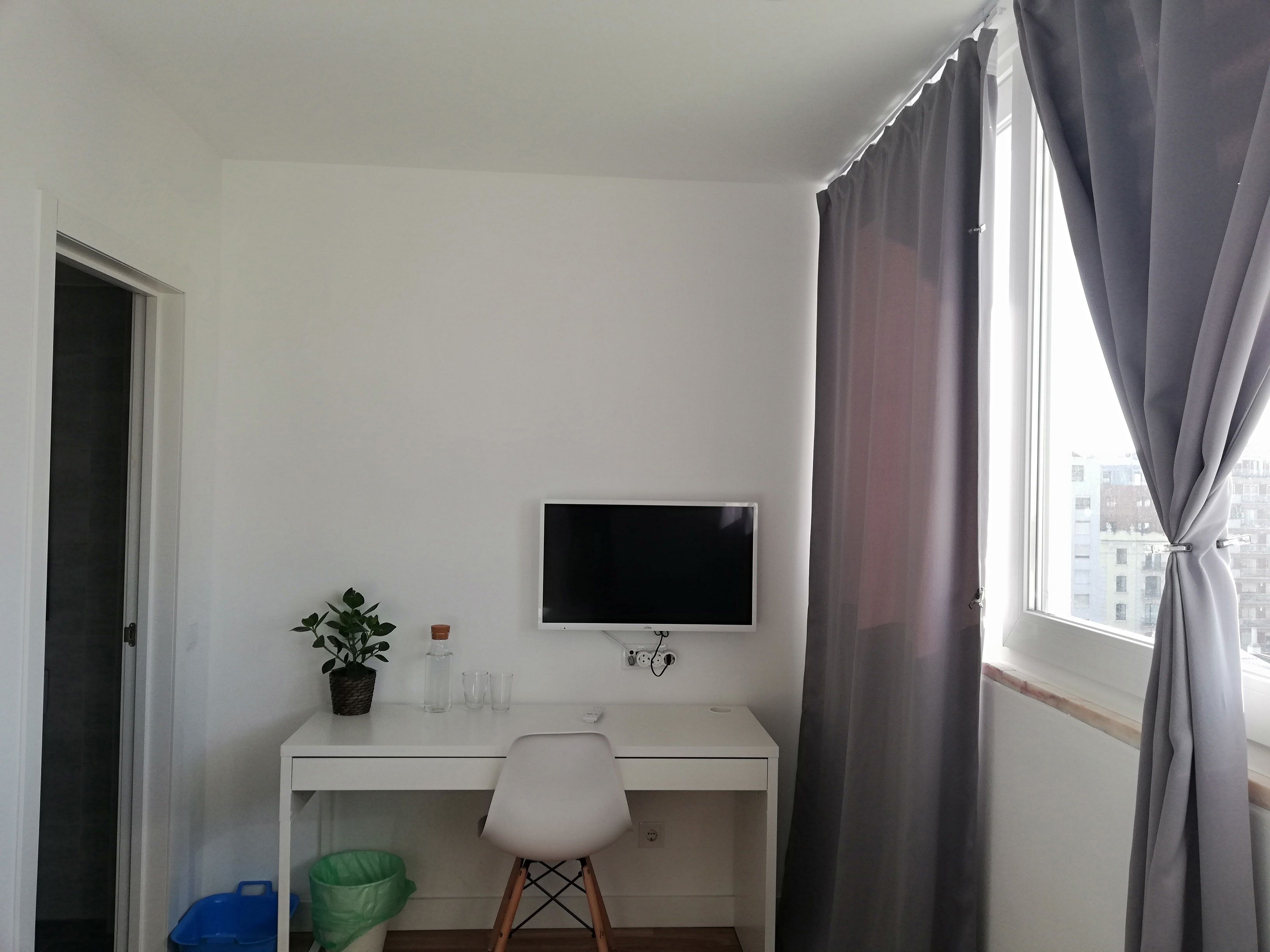 A small, white room with a desk, a chair, a TV, a potted plant, and a window with curtains.