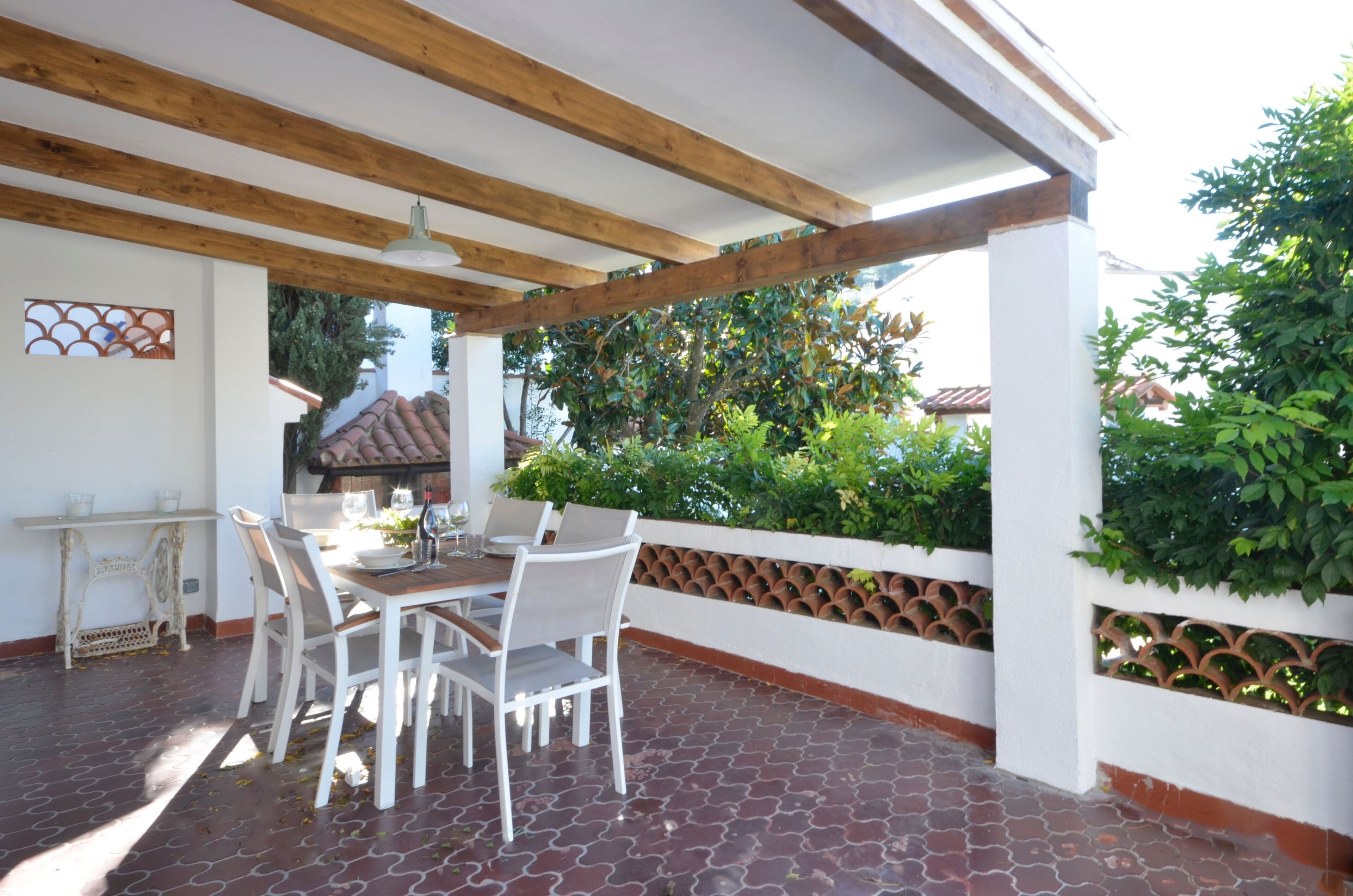 A covered patio with a table and chairs, a tiled floor, and a view of greenery and a building in the background.