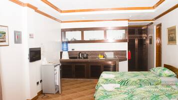 A compact hotel room with a kitchenette, a bed with a tropical-themed bedspread, and a small desk.