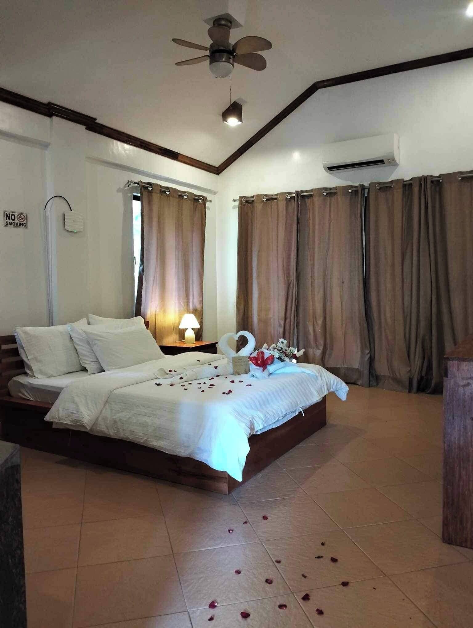 A bedroom with a bed, curtains, a ceiling fan, an air conditioning unit, and a bedside table with a lamp.