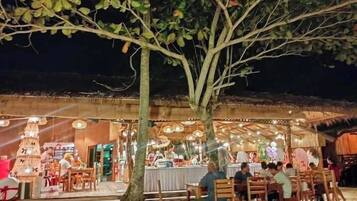 Zona pranzo all'aperto con tavoli e sedie, una struttura con tetto di paglia e un albero sullo sfondo.