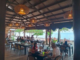Un área de comedor al aire libre con mesas y sillas, techo de paja y vista al mar.