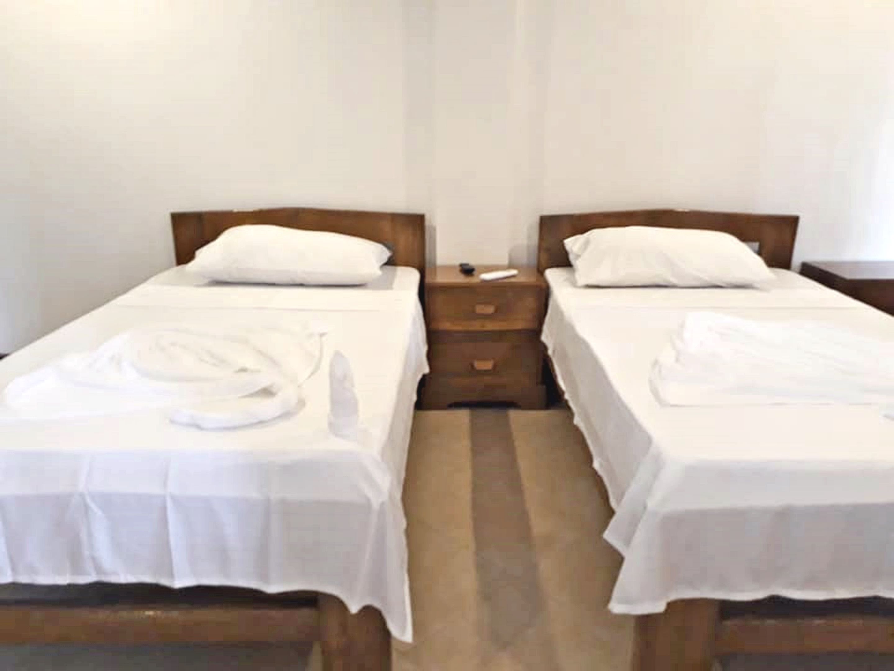 Habitación de hotel con dos camas individuales, cada una con cabecero de madera, ropa de cama blanca y una toalla doblada prolijamente sobre la cama. Hay una mesita de noche de madera entre las camas, con un control remoto encima.