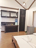 Habitación de hotel con una cama, cabecera de madera, kitchenette pequeña con pileta y dispensador de agua, armario y mueble auxiliar.