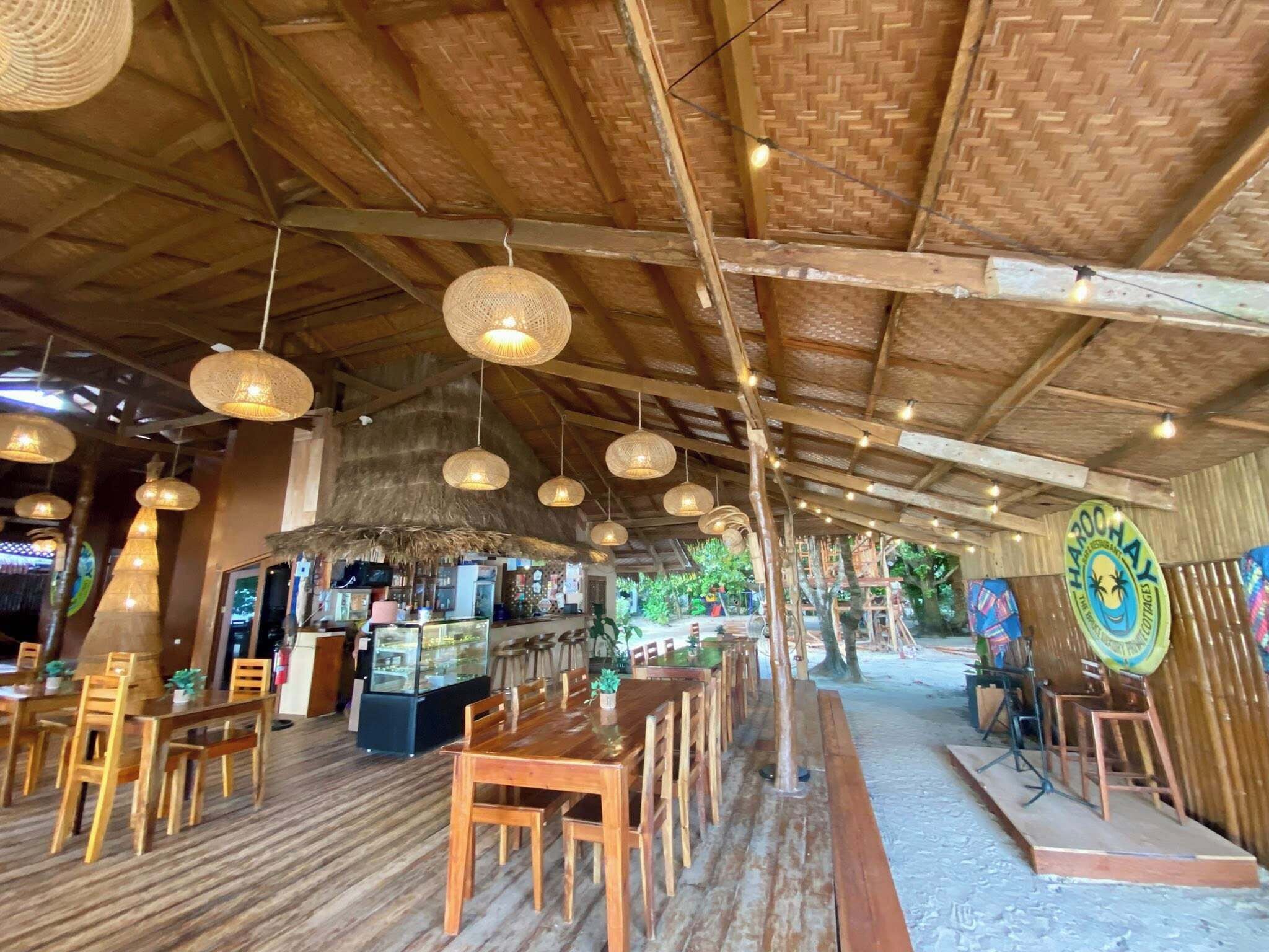 Un restaurant avec des tables et des chaises en bois, des luminaires tressés suspendus au plafond et un toit de chaume.