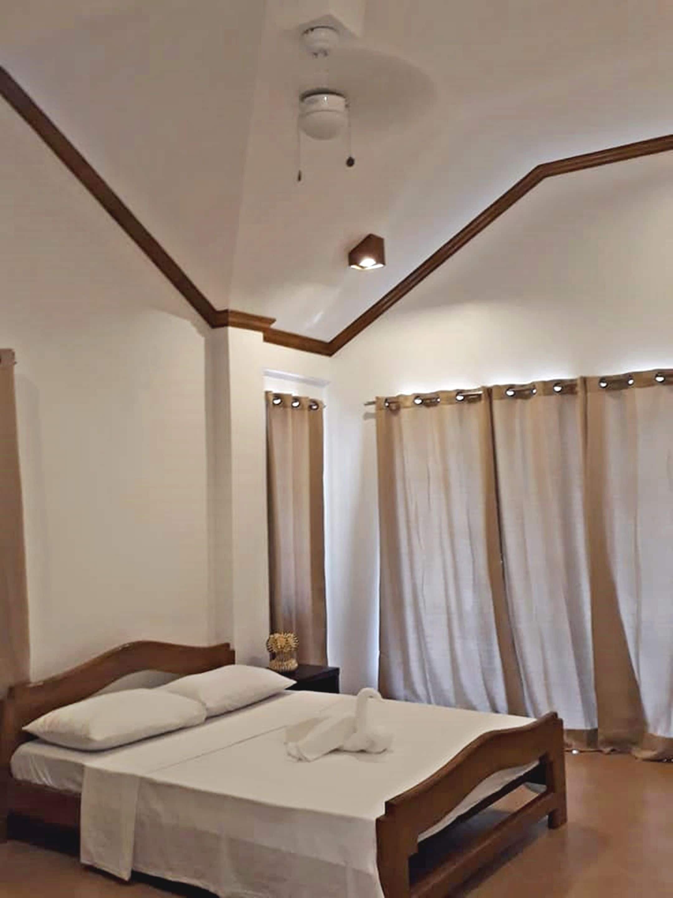 A bedroom with a wooden bed, white bedding, a ceiling fan, and curtains.