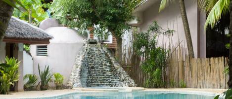 Una piscina con cascada de piedra, rodeada de plantas tropicales y una valla de bambú.