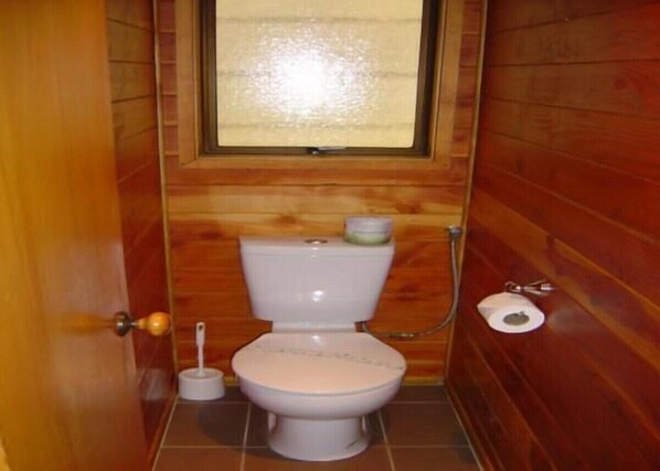A small, wooden-paneled bathroom with a toilet, a window, and a toilet paper holder.