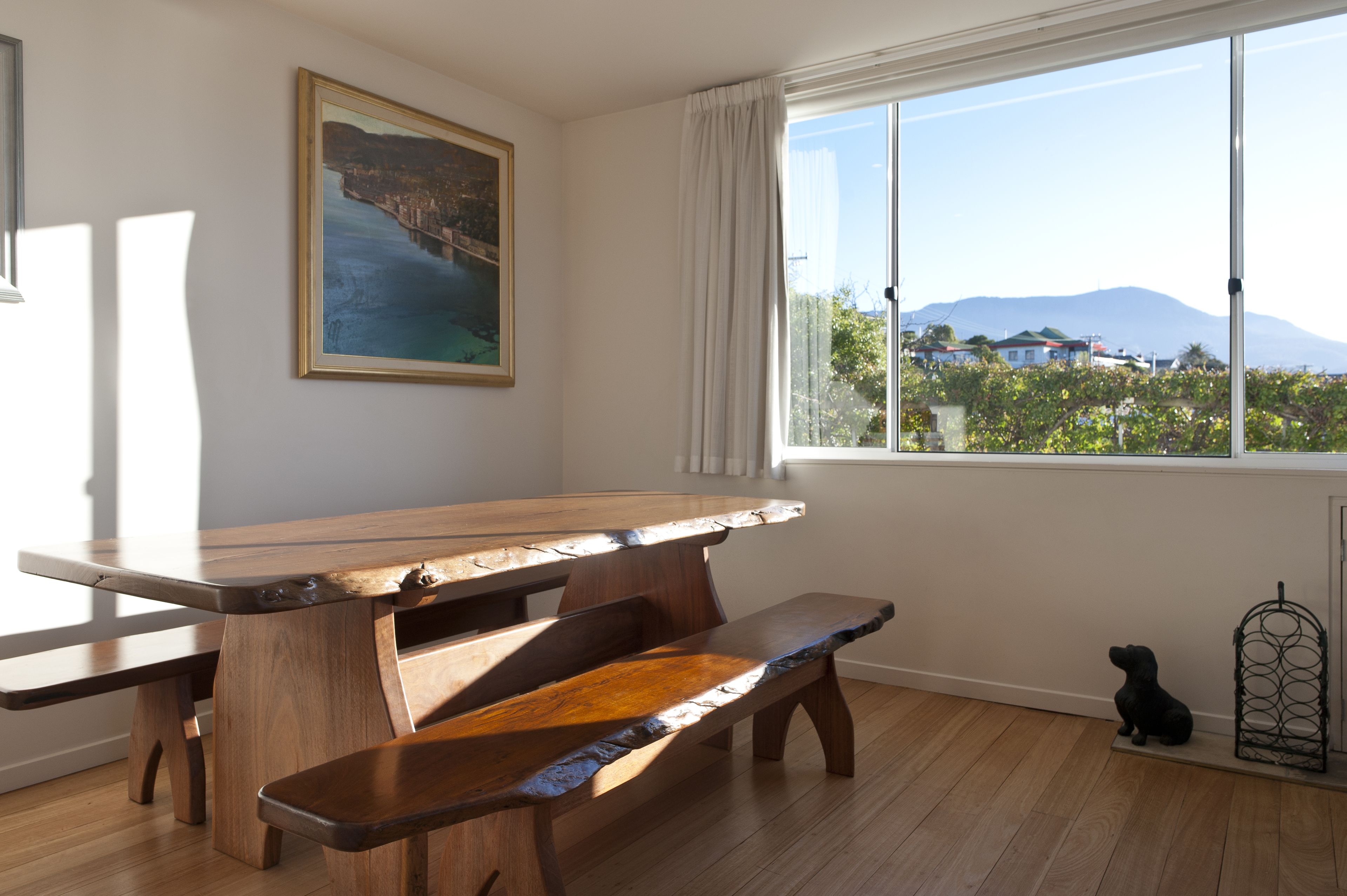 A wooden picnic table with benches, a framed painting on the wall, and a view of houses and mountains through a window.