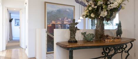 A hallway with a wooden table, a vase of flowers, and a painting of a coastal town.