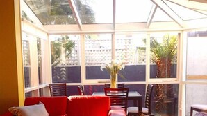 A room with a red sofa, a dining table with chairs, and a large window with a view of greenery and a fence. - Waterloo Cottage (Battery Point)