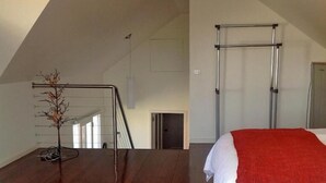 A spacious room with a sloped ceiling, a wooden floor, a bed with a red blanket, and a metal ladder against the wall. - Waterloo Cottage (Battery Point)