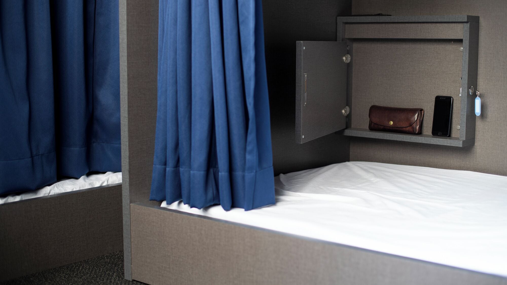 A single bed with a white mattress, a grey headboard, a grey wardrobe with a brown leather item, and a mobile phone on a shelf.