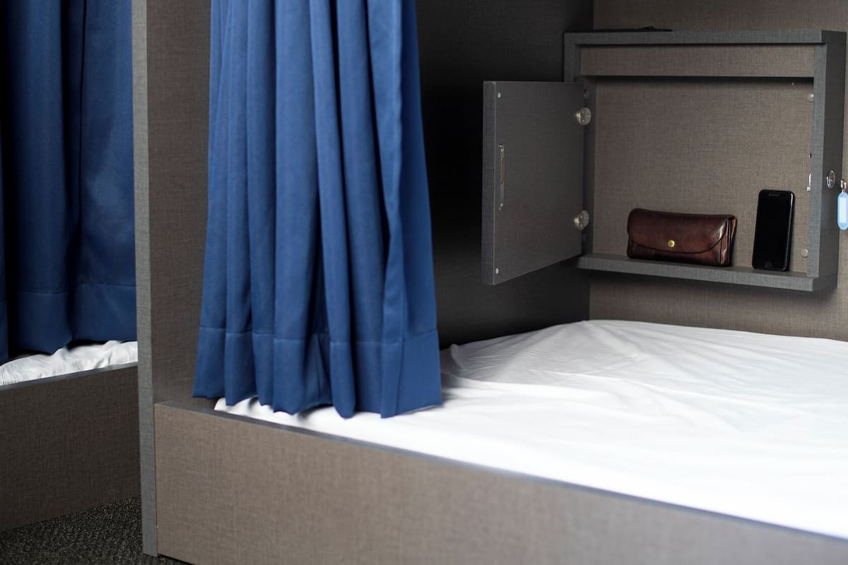 Een enkel bed met een witte matras, een grijs hoofdbord, een grijze garderobe met een bruin lederen voorwerp, en een mobiele telefoon op een plank.