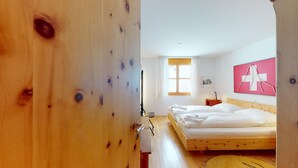 Eine Holztür, die in ein Schlafzimmer führt, mit einem Bett, einem Nachttisch, einem Fenster mit Vorhängen und einer Wanddekoration mit einem roten Kreuz.