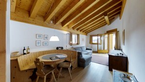 A cozy room with a wooden ceiling, a round wooden table with chairs, a grey sofa, and a small desk with a lamp. - Quiet 2-room apartment, ca. (Sils/Segl Maria)