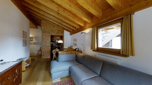 A cozy living room with a gray sectional sofa, wooden ceiling beams, a kitchen area, and a large window with curtains. - Quiet 2-room apartment, ca. (Sils/Segl Maria)