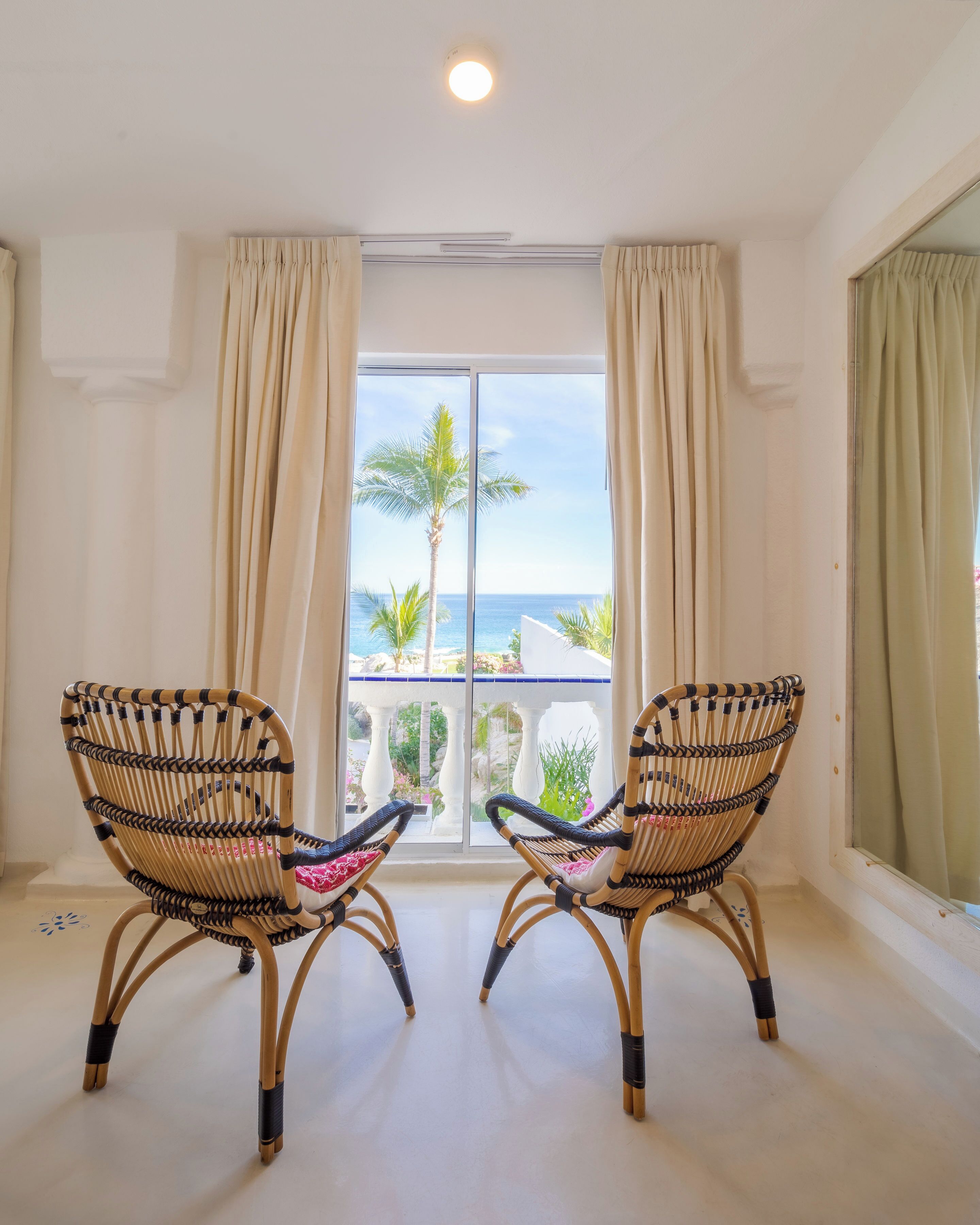 A room with two wicker chairs facing a balcony with a view of the ocean and palm trees.