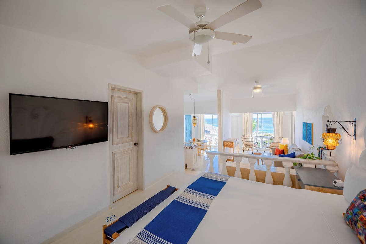 Una habitación de hotel con cama, televisión, ventilador de techo, escritorio con lámpara y vista al océano.