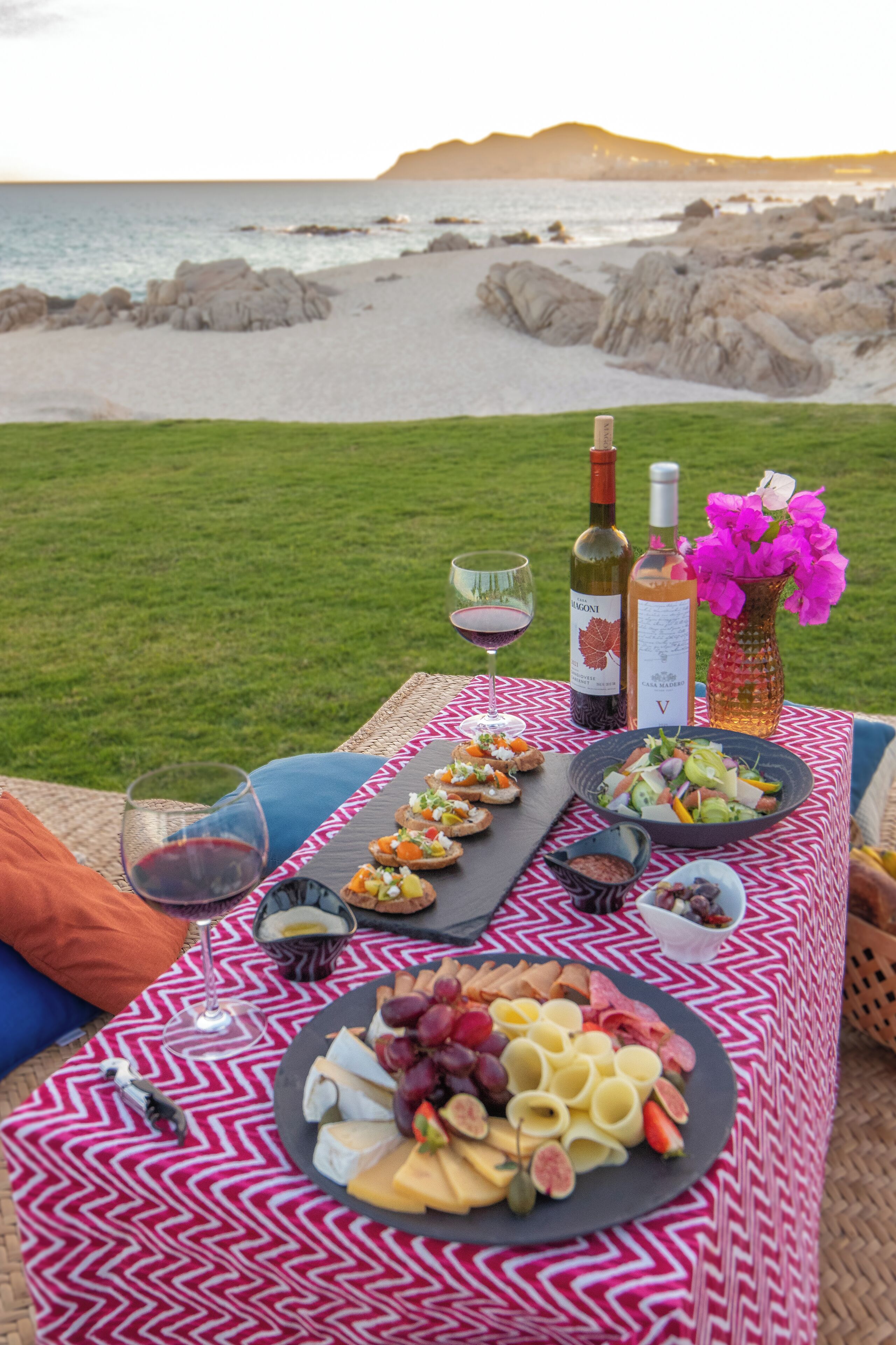 Un montaje para picnic con diversos quesos, frutas y una botella de vino sobre un mantel estampado, con vista a una playa con rocas y un área verde.