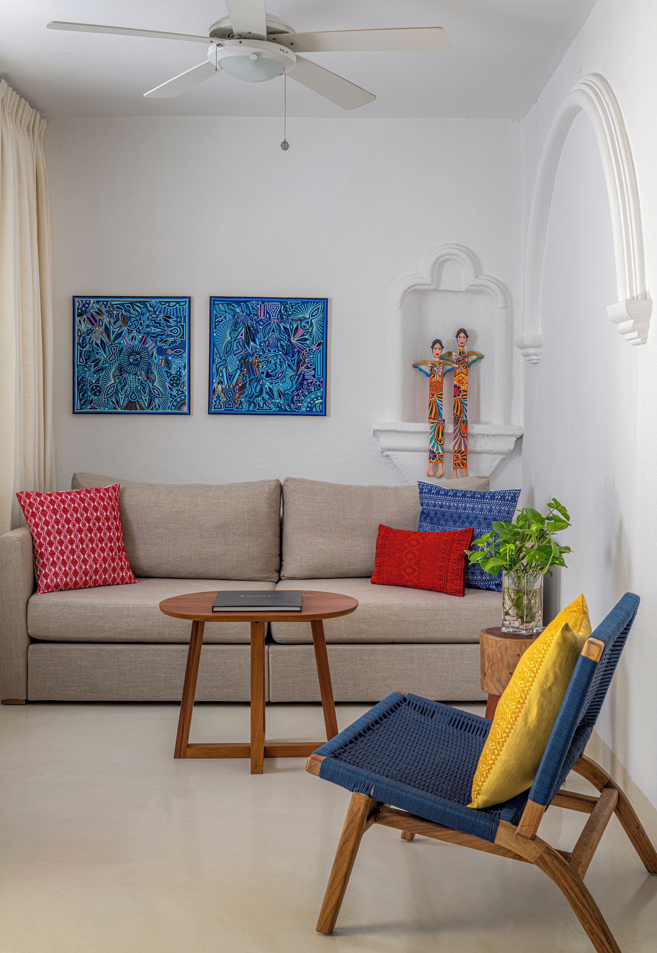 Una sala de estar con un sofá, una mesa de centro de madera, una silla azul tejida y dos cuadros abstractos en la pared.