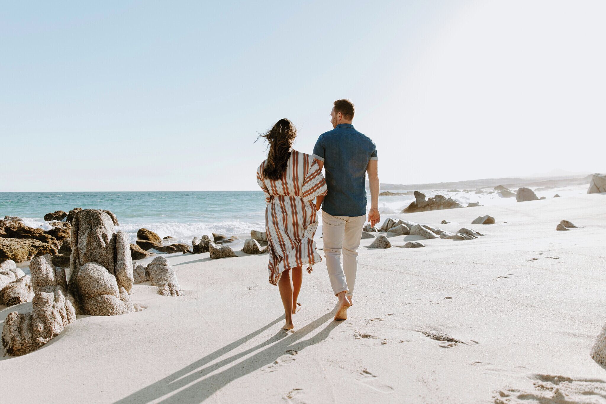 A couple walking on a sandy beach with large rocks and the ocean in the background.