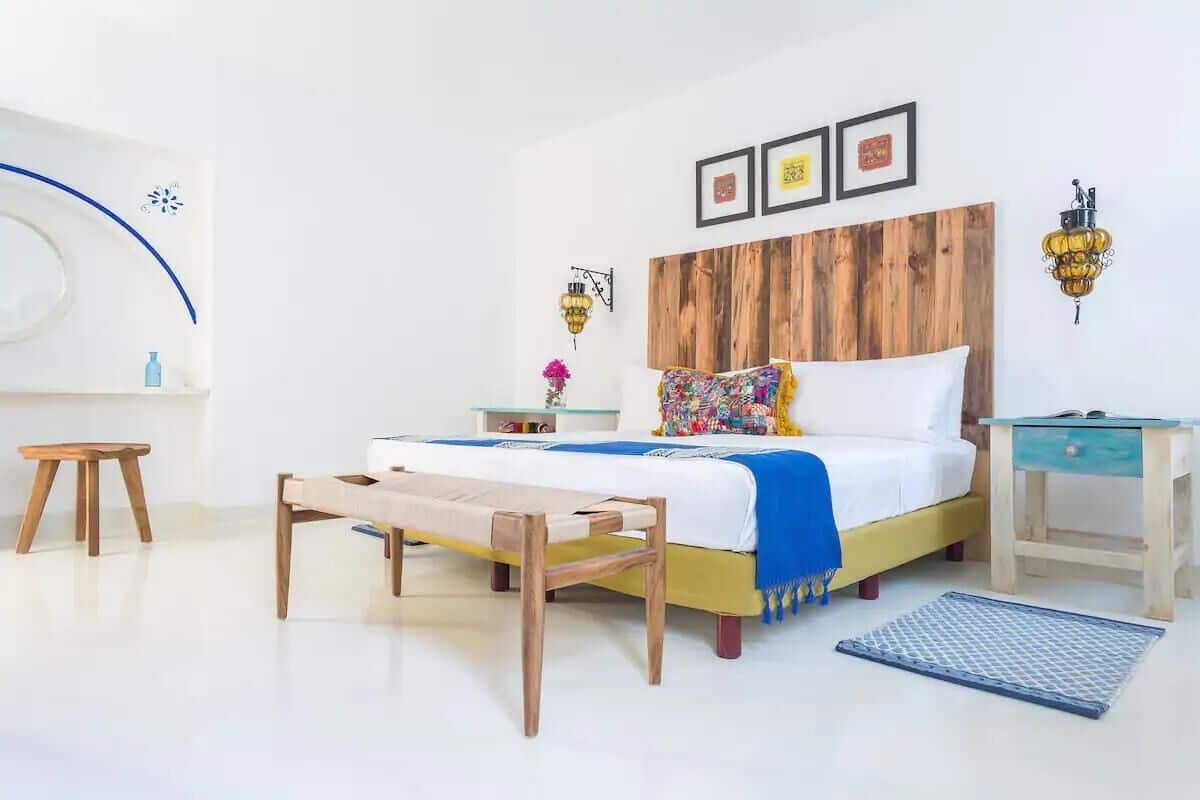 Een slaapkamer met een houten hoofdbord, een bed met een kleurrijk kussen, een nachtkastje, een klein krukje en een wandlamp.