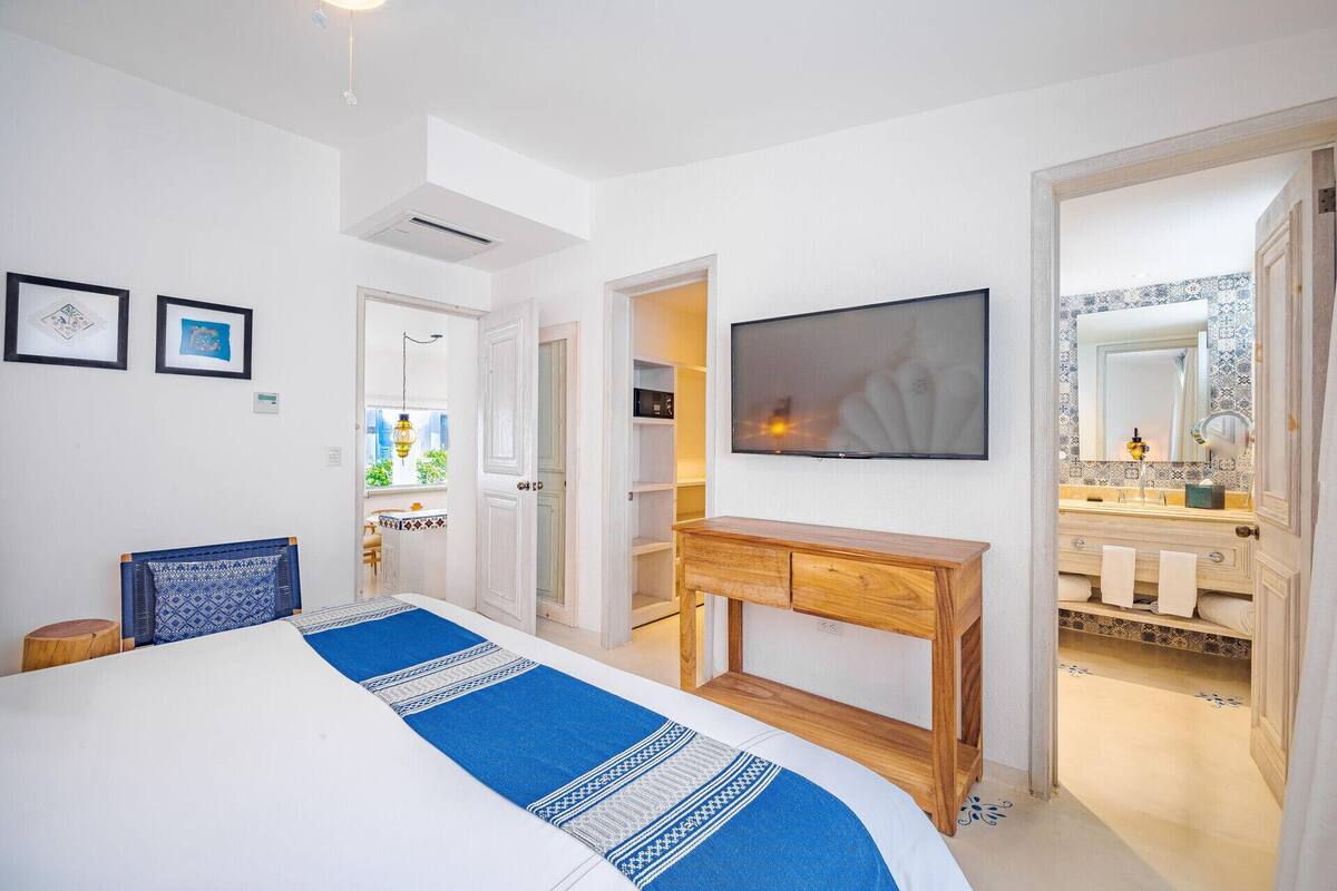 Een slaapkamer met een bed, een televisie, een houten console en een badkamer met spiegel en wastafel.