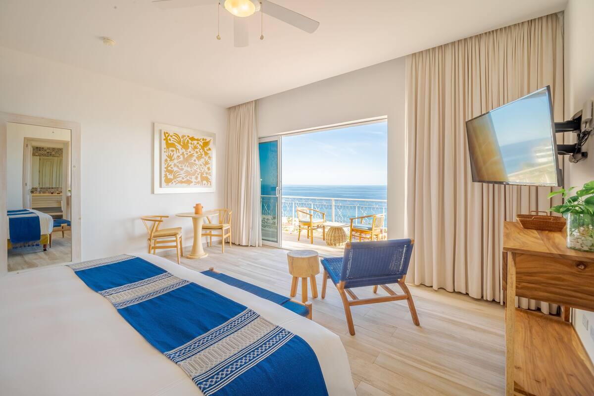 Een hotelkamer met een groot bed, een balkon met uitzicht op zee, een bureau met een televisie en een kleine tafel met stoelen.
