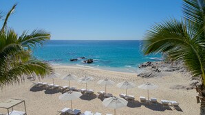 A beach with white sun loungers and umbrellas, a clear blue sea, and rocky outcrops.