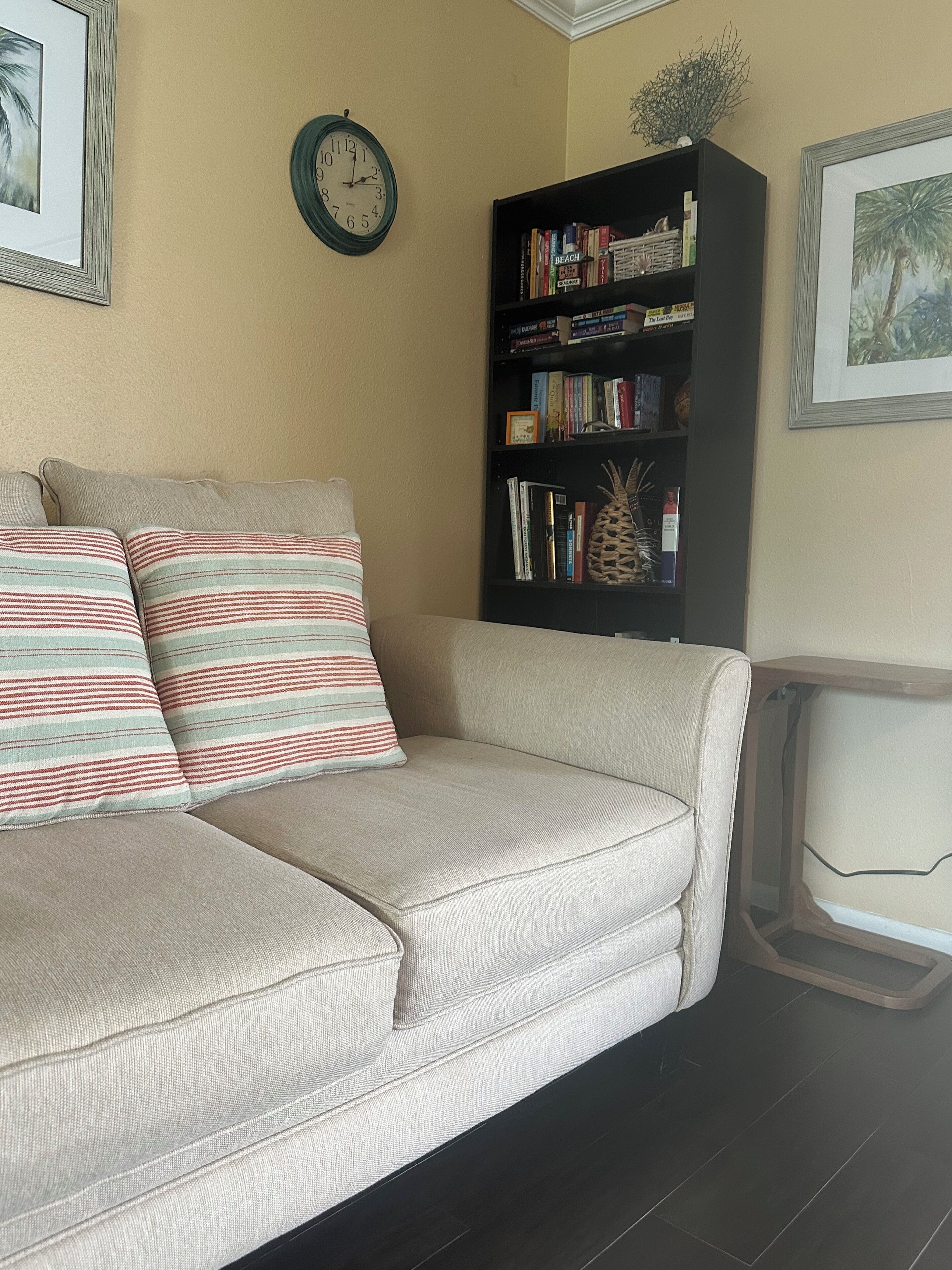Uma sala de estar com sofá bege, almofadas listradas, estante de livros, relógio e obras de arte emolduradas nas paredes.