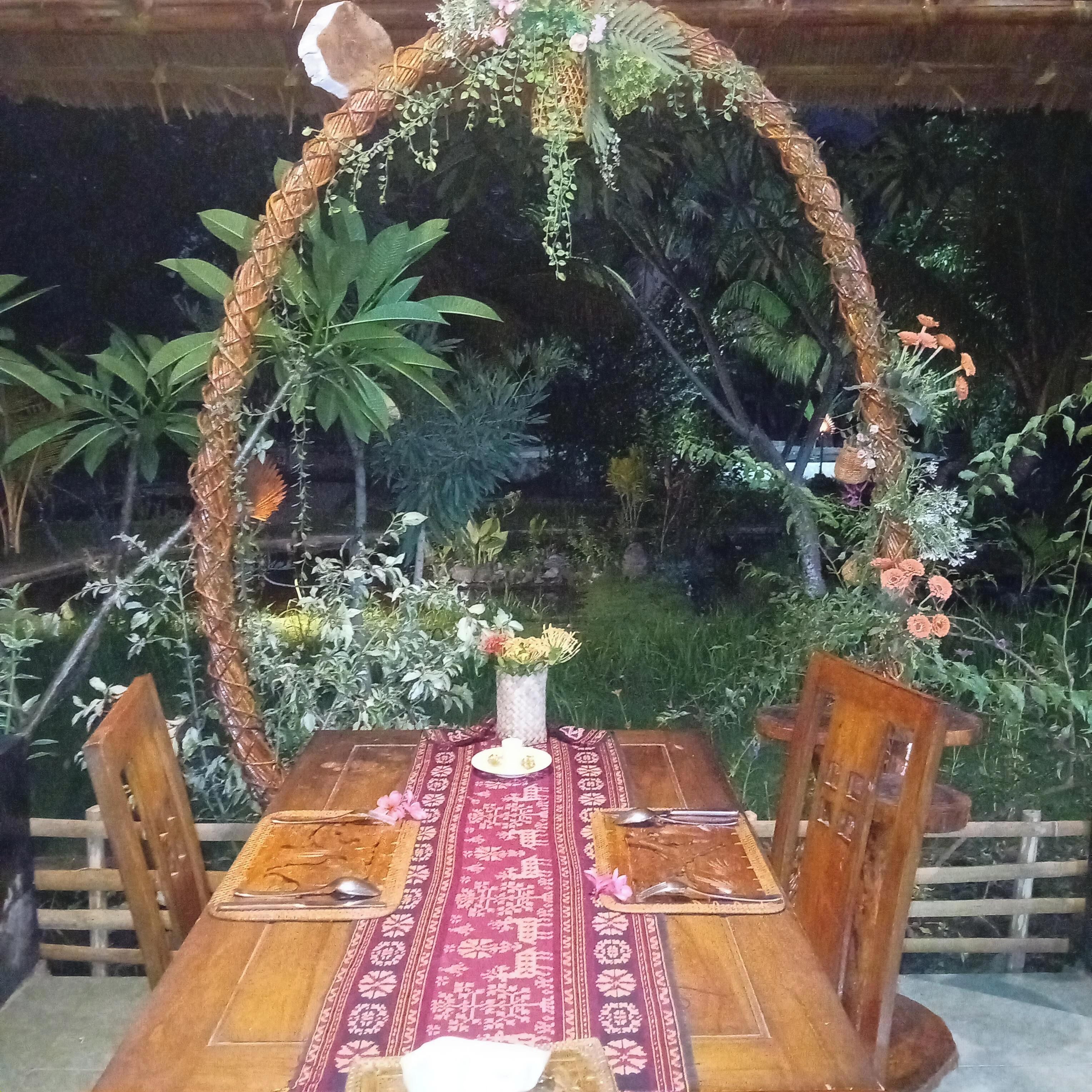 A wooden dining table with a red patterned tablecloth, surrounded by wooden chairs. A decorative arch made of natural materials and greenery frames the table.