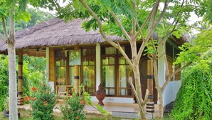 A traditional thatched-roof house with a porch, surrounded by lush greenery and flowering plants. - Budi Sun Resort (Maumere)
