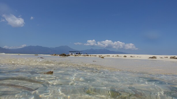 A beach with clear water, distant mountains, and a vehicle on the sand. - Budi Sun Resort (Maumere)