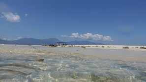 A beach with clear water, distant mountains, and a vehicle on the sand. - Budi Sun Resort (Maumere)