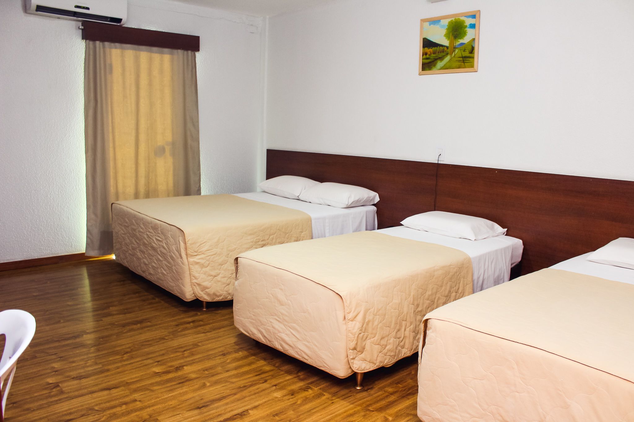 Quarto de hotel com três camas simples, piso de madeira, parede branca com um quadro e uma janela com cortinas.