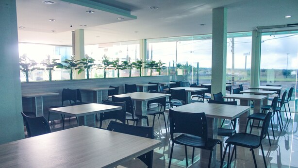 Una cafetería moderna con varias mesas y sillas, ventanas amplias y plantas en macetas.