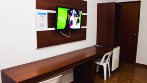 Una habitación con un escritorio de madera, un televisor montado en la pared, una silla blanca y una puerta cerrada.