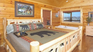 Un dormitorio de cabaña de madera con una cama decorada con almohadas con motivos de osos, un cuadro colgado de osos, una ventana con vista a la ciudad y una mesita de noche de madera con una lámpara.