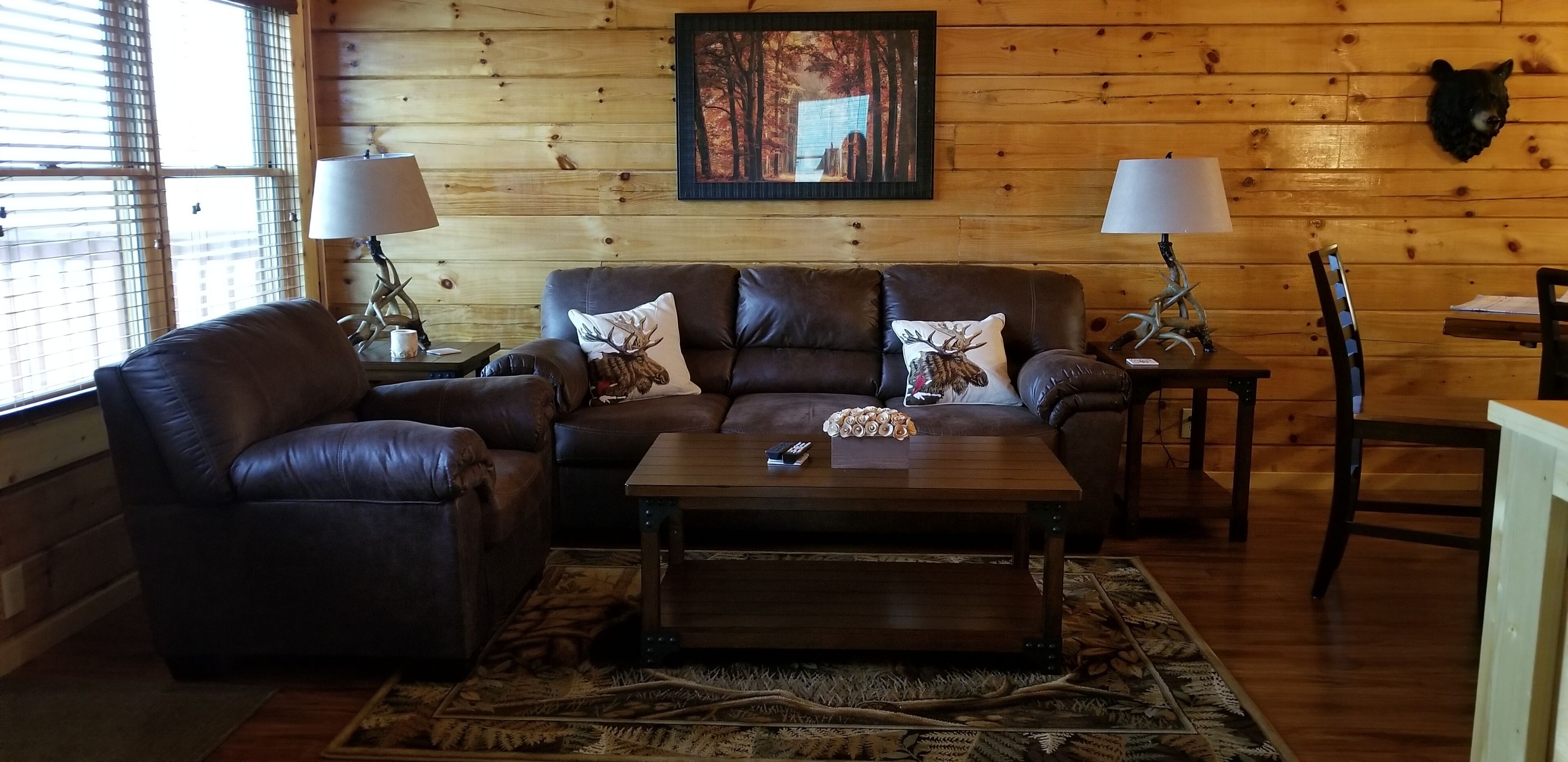 A cozy living room with a leather sofa, two armchairs, a wooden coffee table, and a rug. There are two lamps on side tables, a framed picture on the wall, and a mounted animal head.
