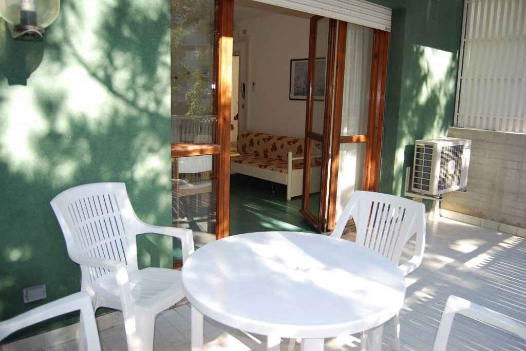 Un área de estar al aire libre con una mesa redonda y dos sillas blancas de plástico, una puerta corredera de vidrio que da a una habitación con cama y un equipo de aire acondicionado a la derecha.