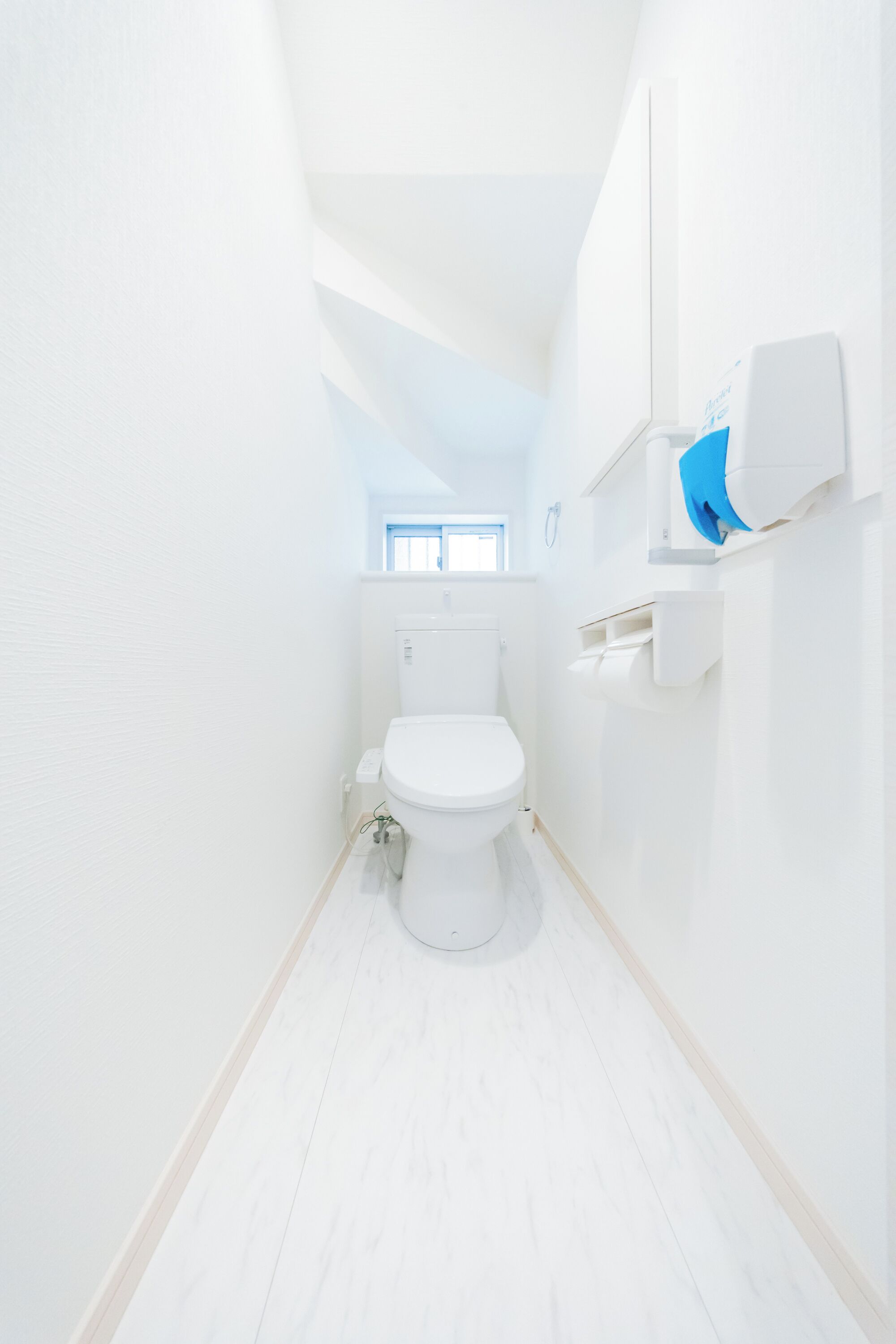 Et aflangt badeværelse med hvide vægge, toilet, et vindue og en hvid hylde med en blå genstand.