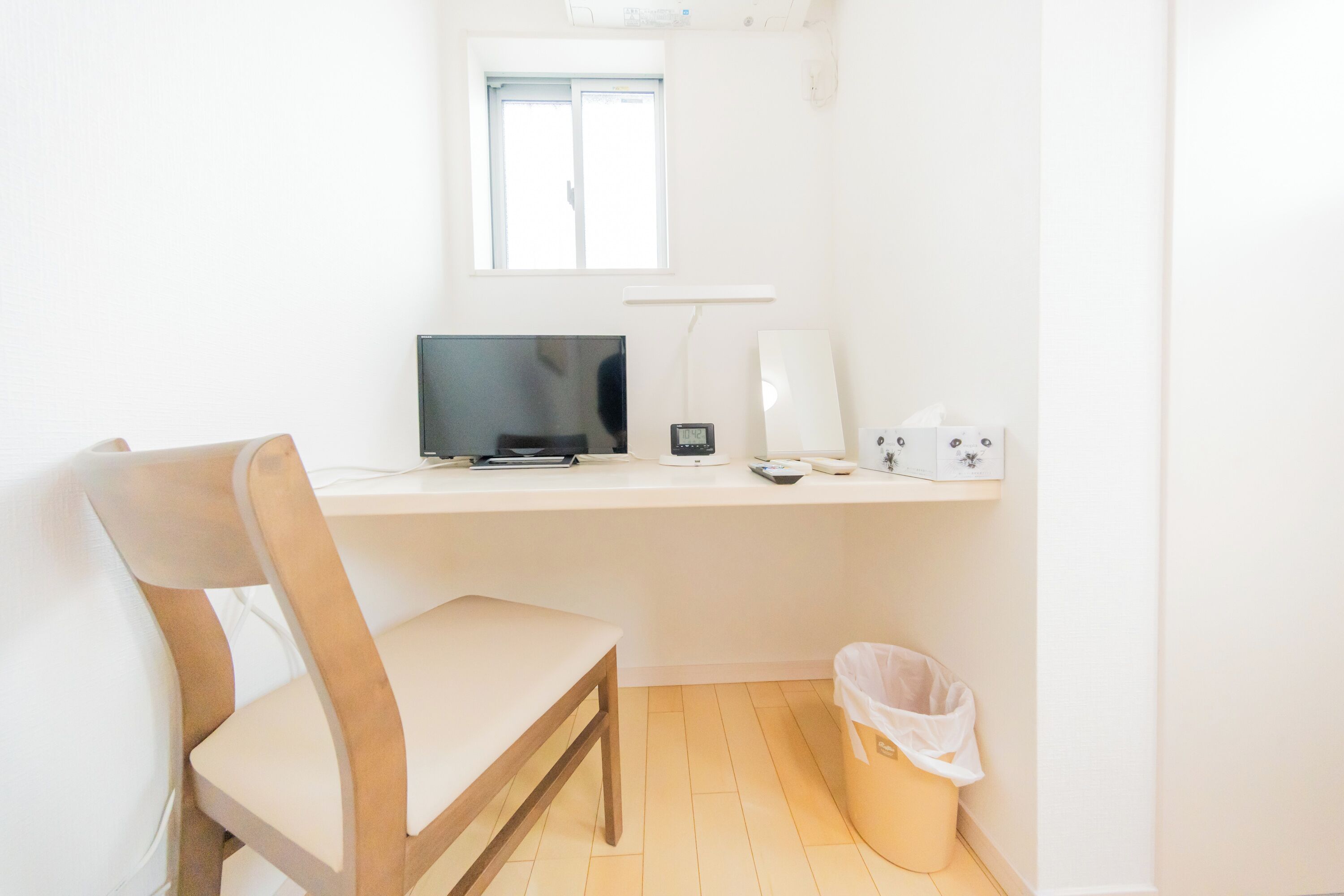 Una habitación pequeña y luminosa con escritorio, silla, computadora, lámpara y un cesto de basura.