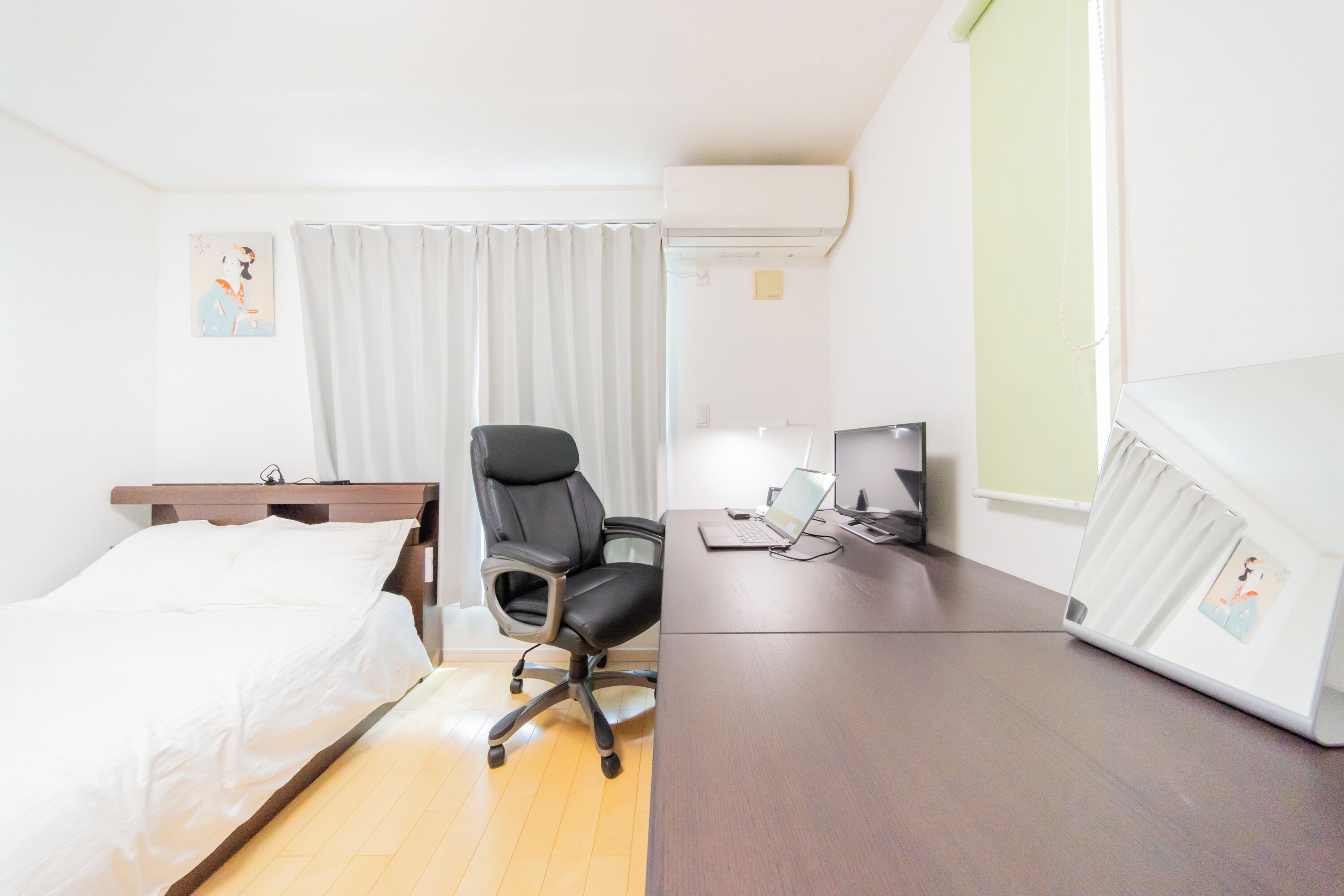 A hotel room with a bed, a desk with a laptop, a chair, and an air conditioning unit.