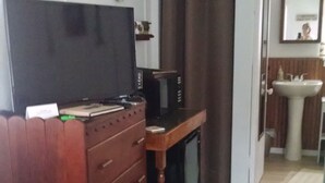 A room with a wooden dresser, a desk with a computer, a television, a bathroom with a sink, and a green carpet. - Eagle Harbor- Harbor Pines: Savor History and Nature at Harbor Pines Cottage (Eagle Harbor)
