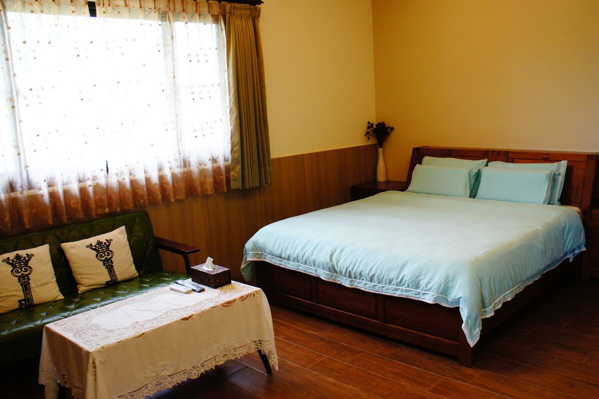 Une chambre à coucher avec un lit en bois, un canapé vert, une petite table recouverte d’une nappe en dentelle et une fenêtre avec des rideaux.