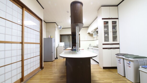 A kitchen with a central island, a refrigerator, cabinets, and a trash bin. - Hikari No Yado (Anan)
