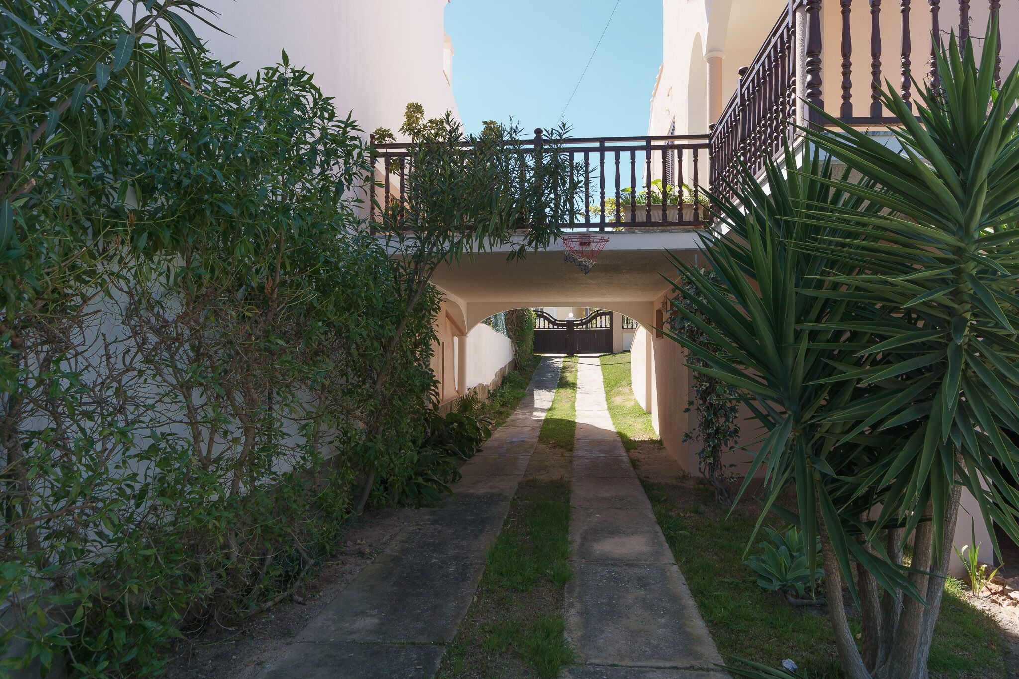 A narrow pathway leading to a gated entrance, flanked by lush greenery and a small bridge with a metal railing.